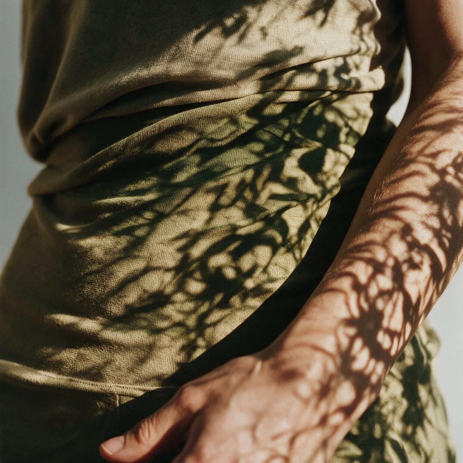 Torso de pessoa em roupa verde com sombras de folhas projetadas, mão sobre o abdômen